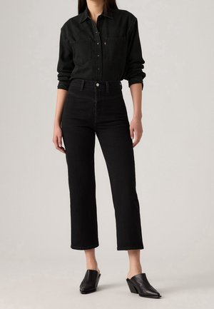 Kobieta ubrana w czarną koszulę na guziki, czarne, krótkie jeansy z wysokim stanem i czarne mules na obcasie, stojąca na jednolitym szarym tle.