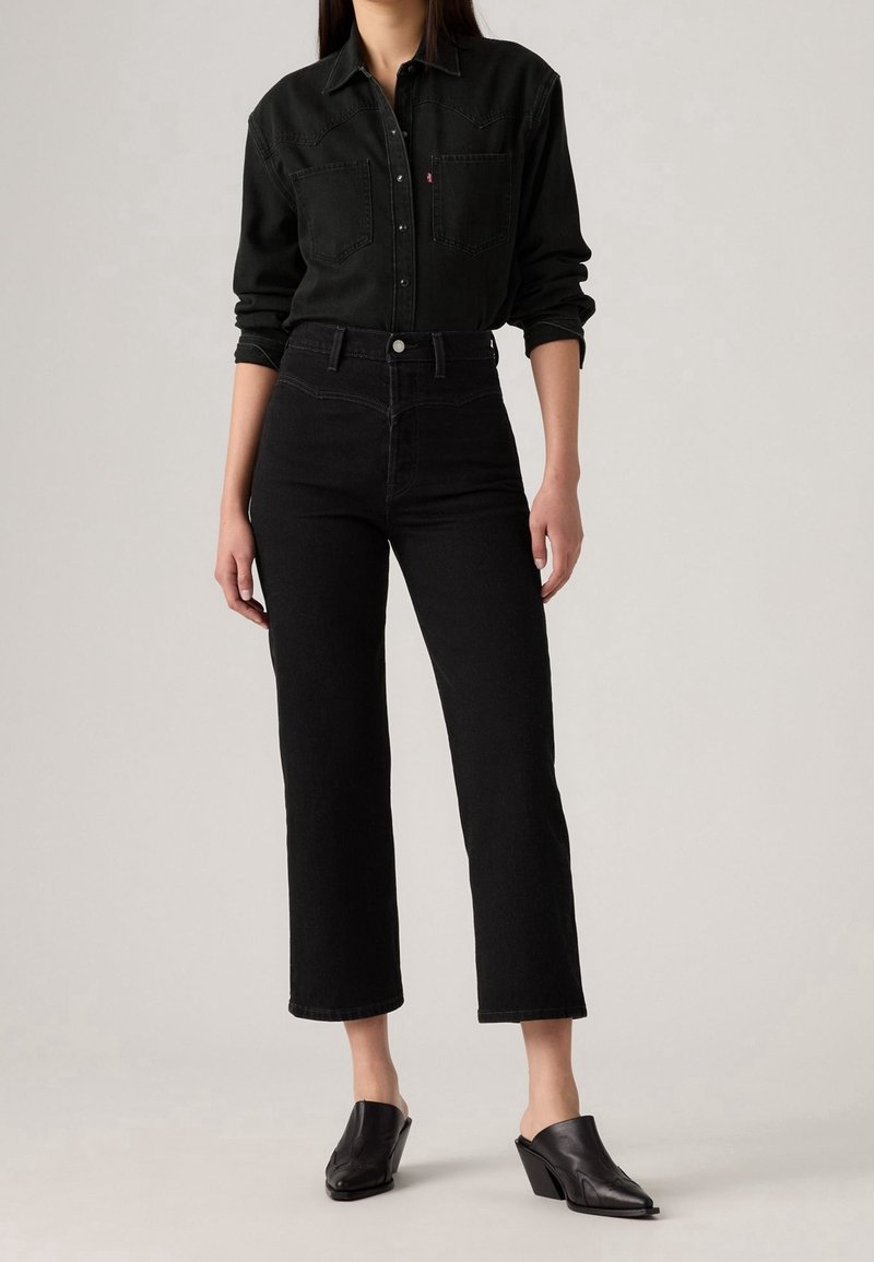 Femme portant une chemise noire boutonnée, un jean noir taille haute coupé au-dessus de la cheville, et des mules noires à talons, debout devant un fond gris uni.