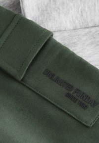 Grünes Stoffpatch mit gesteppter Kante, geprägt mit dem Text "UNLIMITED FUNDAY" und "SINCE 1982", auf hellem grauem Textil gelegt.