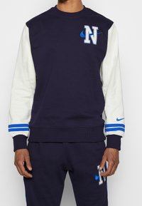 Navy blue sweatshirt with white sleeves, features "N" and Nike logos. Ribbed cuffs with blue stripes and a round neckline. Soft fabric.
