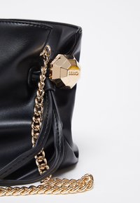 Sac seau en cuir noir avec bandoulière en chaîne dorée et un charme géométrique doré gravé de "Liu Jo" sur fond blanc.