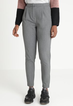 Osoba ubrana w dopasowane szare spodnie i czarne trampki, stojąca z rękami swobodnie opuszczonymi wzdłuż ciała na jednolitym tle.