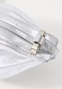 Borsa a mano metallica argento con texture liscia, caratterizzata da cuciture bianche e da una chiusura in metallo argento prominente. Nome del marchio impresso sul lato.