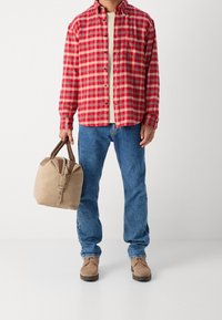 Czerwona i beżowa koszula w kratę, kremowa koszulka, niebieskie dżinsy i beżowa torebka z zamszu. Brązowe buty z płaską podeszwą, lekką teksturą i zaokrąglonym kształtem.