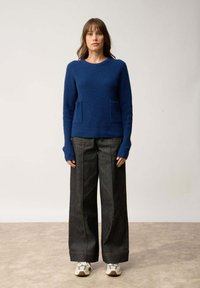 Pull en maille côtelée bleu avec un col rond et deux poches avant, associé à un pantalon en denim noir à jambes larges et des baskets grises.