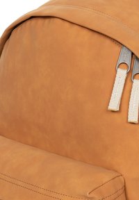 Rugzak van tan leer met een gladde textuur, voorzien van twee zilverkleurige ritssluitingen en crèmekleurige stoffen accenten. Eenvoudig, minimalistisch ontwerp.