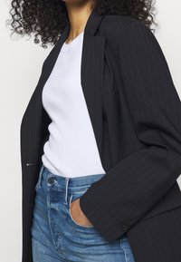 Blazer negro de rayas con un corte relajado, llevado sobre una camiseta blanca ajustada. Jeans de mezclilla azul, mostrando un estilo casual pero pulido.