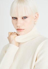 Svijetlosmeđa turtleneck pulover s rebrastim manžetama, mekane teksture. Model drži ovratnik, ističući njegov dizajn visokog vrata i minimalistički stil.