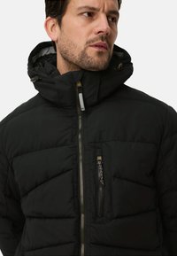 Czarna pikowana kurtka z wysokim kołnierzem, przednim zamkiem błyskawicznym i wieloma kieszeniami. Charakteryzuje się pikowaną fakturą i kontrastującymi elementami zamka.