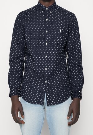 Mann trägt ein dunkelblaues Hemd mit kleinen weißen Mustern und hellblaue Jeans, steht vor einem schlichten Hintergrund.