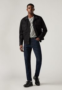 Joven de pie vistiendo una chaqueta de mezclilla negra, camiseta gris claro, jeans oscuros y zapatos de cuero negros frente a un fondo liso.