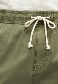 Κοντινό πλάνο σε ελιά πράσινο παντελόνι με ελαστική μέση και λευκό κορδόνι δεμένο σε φιόγκο πάνω από τη ραφή της πλαϊνής τσέπης.