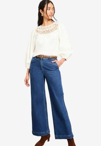 Chemise blanche avec des détails en dentelle au niveau du cou, associée à un jean bleu à taille haute et à jambes larges, et une ceinture marron. Des bottes à talons marron complètent le look.