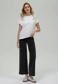 T-shirt branca de manga curta com bordados florais nos ombros, combinada com calças largas pretas e sapatilhas prateadas.