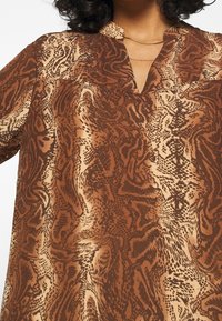 Braun-beige gemusterte Bluse mit lockerer Passform, die einen leichten V-Ausschnitt und einen strukturierten Stoff aufweist, der eine fließende Silhouette zur Schau stellt.