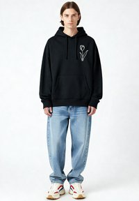 Sweat à capuche noir avec une poche kangourou à l'avant et un motif tulipe blanche sur la poitrine, associé à un jean large bleu clair et des baskets blanches.