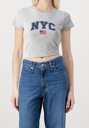 Kvinde iført en grå cropped top med teksten ""NYC"" og et amerikansk flag over højtaljede blå jeans mod en ensfarvet baggrund.