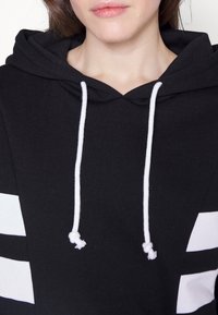 Gros plan sur une personne portant un sweat à capuche noir avec des cordons blancs et des bandes blanches rectangulaires sur les manches.