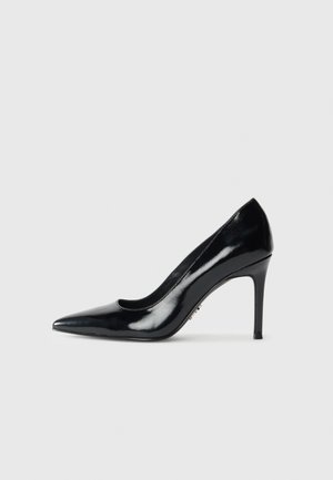 Schwarzer Lackleder-Pumps mit spitzer Spitze und schmalem Stiletto-Absatz auf schlichtem hellgrauem Hintergrund.