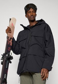 Black ski jacket with multiple front pockets, adjustable hood, and textured fabric. Model holding brown skis with bindings.