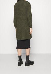 Lång olivgrön stickad cardigan med texturerade ärmar, i kombination med en svart ribbad kjol och svarta snörkängor. Bakifrån vy av outfiten.