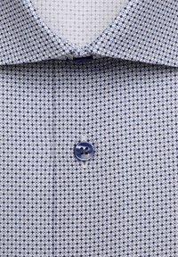 Chemise blanche avec un petit motif carré bleu marine, avec un bouton bleu et un col classique.