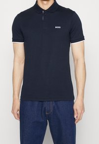 Man som bär en marinblå polo med "BOSS"-logotyp på bröstet och mörkblå jeans, står mot en enfärgad bakgrund.