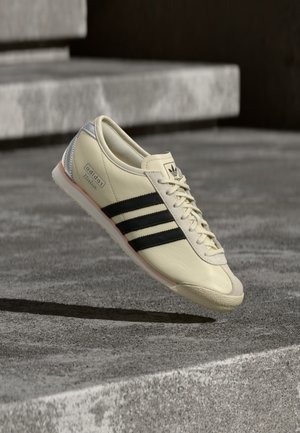 Adidas Italian lenkkari kermaan vivahtavasta nahasta, mustilla raidoilla, pyöreällä varpaalla ja vaaleanharmailla kantapääyksityiskohdilla tekstuurisella harmaalla pinnalla.