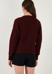 Burgundy knit sweater with a textured pattern, ribbed hem, and long sleeves, viewed from the back. Paired with black shorts.