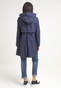 Veste à capuche imperméable bleu marine avec une taille cintrée, un dos ventilé et un ourlet plus long. Portée avec un jean aux revers retroussés et des baskets à motifs.