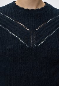 Gros plan d'un pull tricoté bleu marine foncé avec un motif texturé et deux rangées diagonales de petits trous formant un V près de l'encolure.