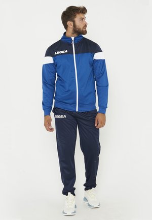 Homme portant une veste de sport bleue et marine ainsi qu’un pantalon avec des baskets blanches, debout et regardant sur le côté sur un fond uni.