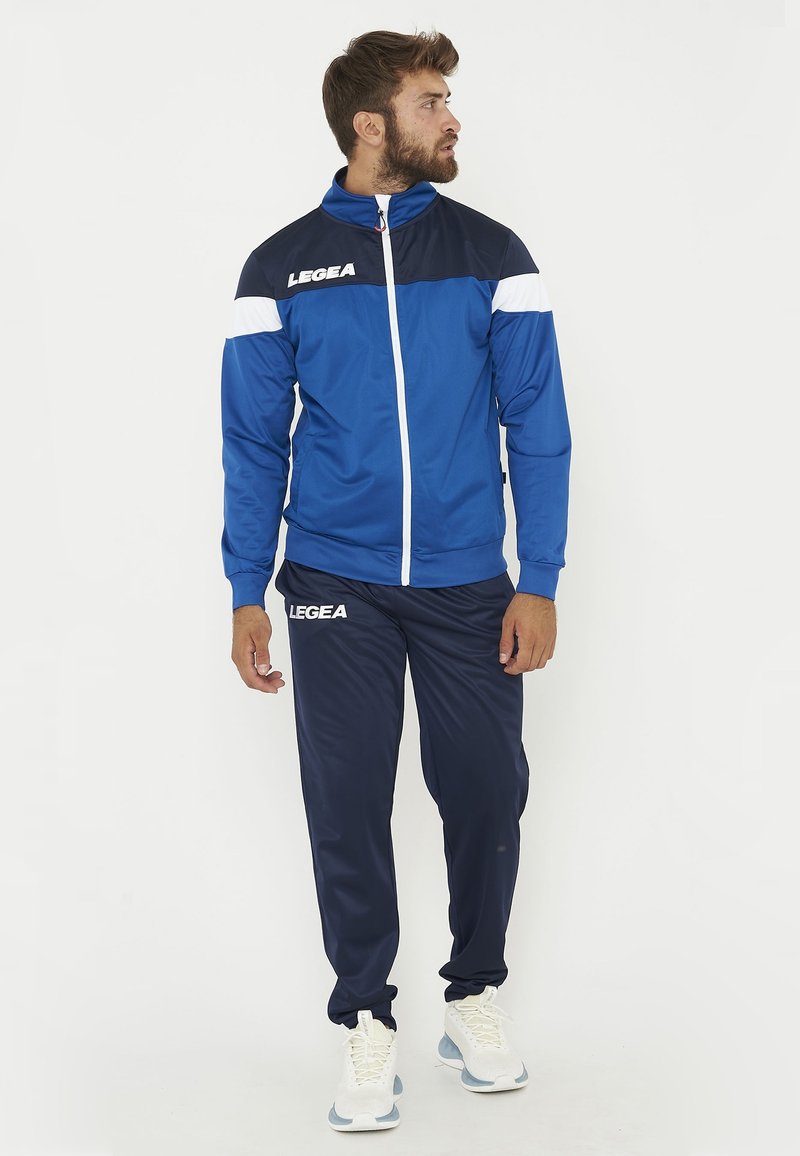 Homme portant une veste de sport bleue et marine ainsi qu’un pantalon avec des baskets blanches, debout et regardant sur le côté sur un fond uni.