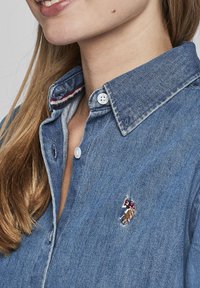 Denim shirt with a button-down collar, light blue color, featuring a small embroidered design on the left chest and contrasting inner collar.