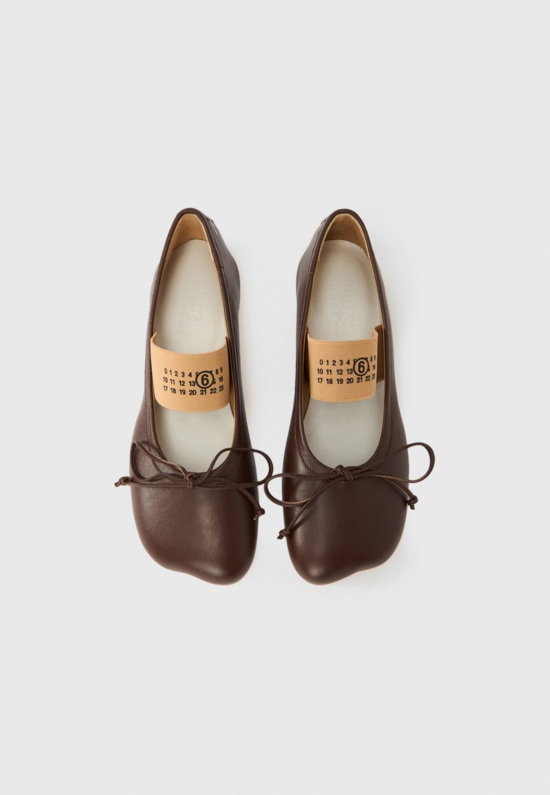 Par af mørkebrune læderballet flats med beige elastikbånd med sorte tal, bundne snørebånd, på hvid baggrund.