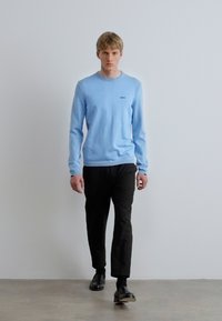 Világoskék, hosszú ujjú pulóver bordázott nyakkivágással, fekete nadrággal és cipővel viselve. Egyszerű kialakítás, minimális márkajelzés a mellkason.