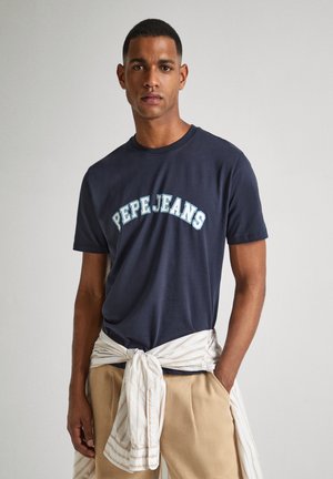 Camiseta de algodón azul marino con "PEPE JEANS" en letras curvas de color azul claro. El modelo también lleva pantalones beige y una camisa a rayas atada a la cintura.