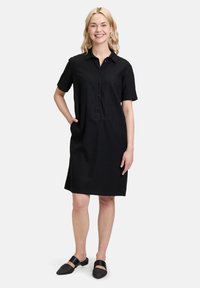 Czarna sukienka na guziki z krótkimi rękawami i bocznymi kieszeniami, wykonana z lekkiego materiału. Modelka nosi pasujące czarne buty wsuwane.