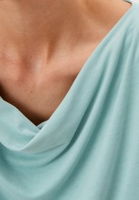 Felpa a collo alto di colore azzurro chiaro realizzata in tessuto morbido, con un design drappeggiato che mette in risalto il décolleté e le clavicole. Texture liscia.