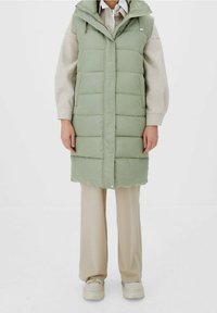 Lichtgroene pufferbodywarmer met een capuchon, voorzien van horizontale doorgestikte naden en drukknopen. Draagt over een beige trui en crèmekleurige broek.