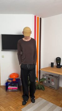 Oseba nosi bež kapo, rjavo dolg rokav majico in črne hlače ter stoji v sobi z oranžno črtasto steno, televizorjem in mizo z različnimi predmeti.