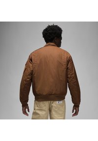 Giubbotto bomber marrone con colletto a costine, polsini e vita elasticizzati. Presenta una tasca a patch sulla manica e un tessuto liscio e dalla texture interessante.
