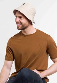 Cappello a secchi beige con una tracolla, abbinato a una camicia a maniche corte marrone con un logo discreto sul petto.