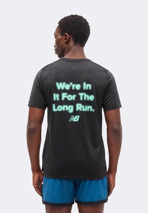 Mand set bagfra iført sort T-shirt med lysende tekst "We're In It For The Long Run" og et New Balance-logo, kombineret med blå shorts.