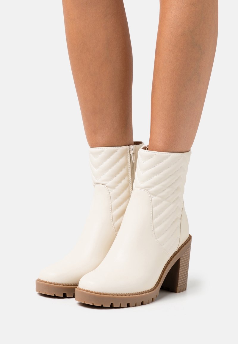 Anklet, joka käyttää kermaista päällystettyä nahkaa nilkkureita, joissa on sivuvetoketjut ja paksut ruskeat kulutusalustat sekä lohkareet valkoista taustaa vasten.