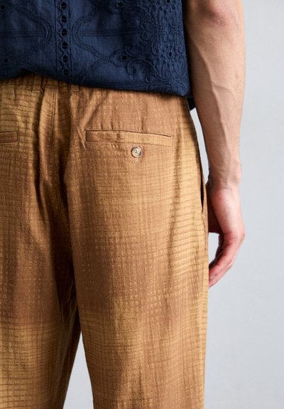 Brown checkered trousers made of textured fabric, featuring two back pockets and a button detail. Worn with a navy embroidered top.