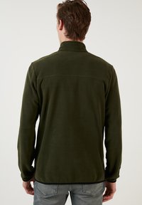 Giacca di pile verde con colletto alto, maniche lunghe e dettagli di cucitura minimi. Tessuto morbido con una vestibilità rilassata, indossata con jeans chiari.