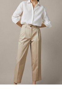 Pantalons beiges ajustés à taille haute, avec des poches latérales et une fermeture à un bouton, associés à une chemise blanche à boutons.