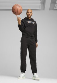 Felpa nera con logo, pantaloni della tuta neri coordinati e sneaker bianche. Tiene un pallone da basket, in piedi su uno sfondo neutro.