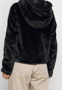 Svart, mjuk fuskpäls hoodie med rund huva, långa ärmar och avslappnad passform. Bärs tillsammans med beiga byxor som visar detaljer i strukturerad väv.